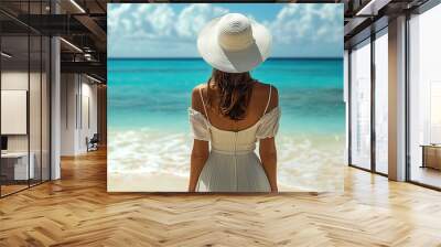 A woman in a dress and white hat standing on the beach, viewed from the back. The turquoise ocean and sky are visible in the background. Wall mural