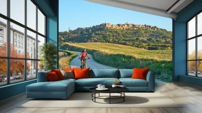 nice senior woman riding her electric mountain bike between olive trees in the Ghianti area with medieval city of Montepulciano in background, Tuscany , Italy Wall mural