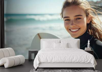 Portrait of a beautiful young european woman in a black wetsuit holding a white surfboard on a beach, smiling at the camera, with an ocean waves and beautiful day with the sun shinning bright Wall mural