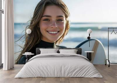 Portrait of a beautiful young european woman in a black wetsuit holding a white surfboard on a beach, smiling at the camera, with an ocean waves and beautiful day with the sun shinning bright Wall mural