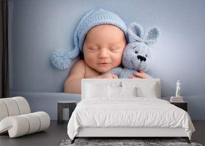 A cute newborn boy in the first days of life sleeps naked on a blue fabric background. The kid gently hugs a light blue knitted bunny. Studio professional macro photography, newborn baby portrait. Wall mural