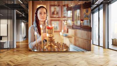 woman in the Russian Orthodox Church with red hair and a scarf on her head lights a candle and prays in front of the icon. Wall mural