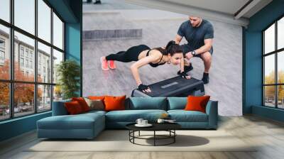 Attractive woman doing push-up with the help of a personal instructor isolated on wooden floor background. Female training with a man coach in fitness center Wall mural