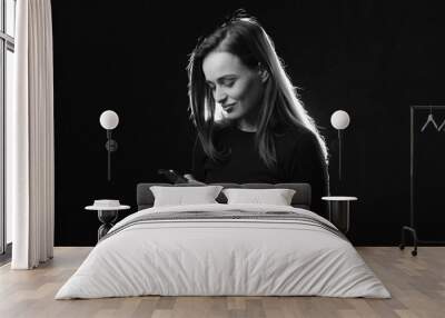 Black and white portrait of young woman looking at phone and smiling, isolated studio photo on dark background. Beautiful model with long hair using cell phone Wall mural