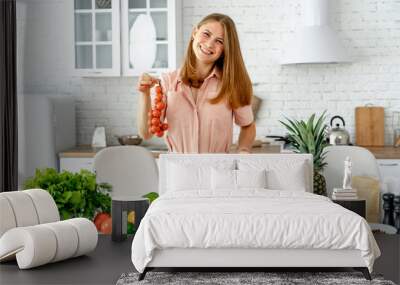 Girl standing at kitchen holding red tomato bunch in her hands. Housewife taking fresh ingredients for vegetable salad. Wall mural