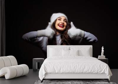 Joyful woman in cozy hat and mittens celebrates winter indoors. A cheerful woman wearing a warm hat and mittens expresses joy while posing in a dark indoor setting during winter. Wall mural