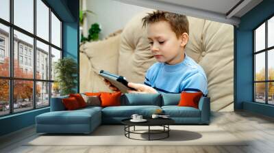 Little good-looking boy sitting on sofa and playing game on digital tablet. Portrait of a young child at home watching cartoon on the laptop. Wall mural