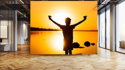 little kid holds two paper planes in spread hands and plays at sunset on the lake background. Time outdoor. Black silhuette against sundown Wall mural