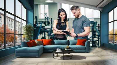 Muscular coach shows the woman's sport results on the background of gym. Personal trainer and young female talking about the progress inside the sports club. Wall mural