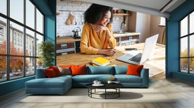 A cheerful mixed-race girl uses laptop for remote work or home leisure while sitting in the kitchen at home. Side view a nice girl with an afro hairstyle looks at screen with a smile Wall mural