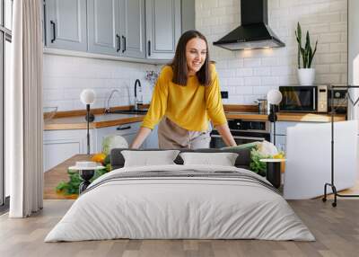 A young woman in yellow shirt cooks in the kitchen and looks with interest at the laptop screen. Woman prepare veggie dish, vegetables on the table Wall mural