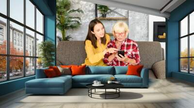 Adult granddaughter teaching the grandmother to use a modern smartphone. Two woman different generations sit on the couch and look at phone screen with interest Wall mural