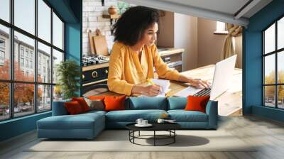 An african-american young woman is using laptop computer for remote work or studying online, she takes notes watching webinars or classes at kitchen table at home Wall mural