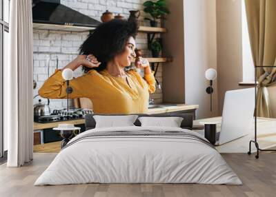 An african-american young woman using laptop computer for remote work or study online at kitchen home, she takes a break and stretch oneself Wall mural