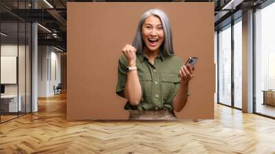 Astonished senior woman holding and using smartphone, mature modern grey-haired lady looking at the camera, receiving unexpected good news, crazy sale deal, isolated over brown background Wall mural