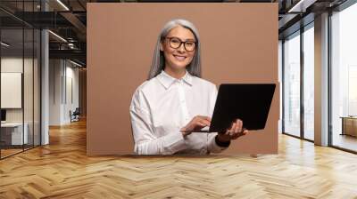 Attractive woman working on new post for her job on laptop, looking at camera with happy inspired smile, watching inspiring video on internet. Indoor studio shot isolated on beige background Wall mural