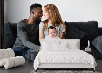 Beautiful caring parents with one child sit one the sofa in living room. Charming husband and pretty wife smiling, cuddling with little daughter. Happy mom and dad enjoy parenting, tenderness concept Wall mural