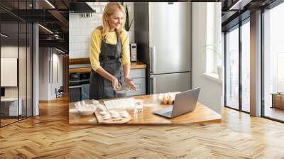 Blonde middle-aged woman cooks at home in the kitchen, housewife in apron watches a video recipe on a laptop and works with the dough on the table Wall mural