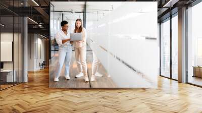 Businesswoman with laptop explaining project details to colleague at office hallway. Business partners having meeting in coworking office space. Two female diverse employees, full length Wall mural