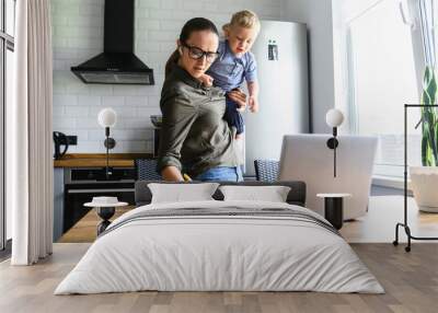 Busy mom works at home with an active toddler on her arms. Mother in glasses is watching on laptop and writing notes in the kitchen while holding kid Wall mural