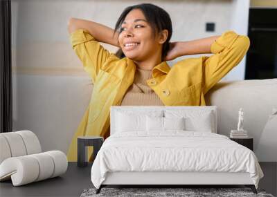 Carefree African-American young woman leaned back on the sofa, holding arms behind head dreaming, serene dark skin lady looks away dreamily, rest, enjoying calm at home Wall mural