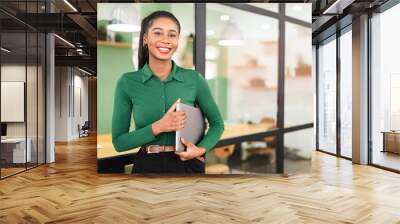 Carefree inspired african-american female employee standing in modern office space and holding laptop, cheerful black businesswoman in green shirt looks at the camera with light friendly smile Wall mural