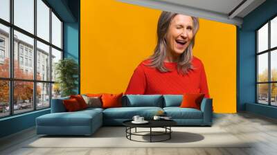 Charming senior woman laughing toothy. Elegant carefree mature modern gray-haired lady in red jumper standing and blinking eye as having cunning idea, winking and flirting to camera. Happy lifestyle Wall mural