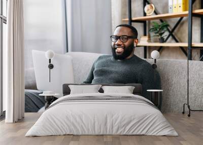 Cheerful African-American guy sits on sofa in modern living room alone and using trendy laptop for net searching, web surfing or texting messages. A black guy in glasses looks into a camera and smiles Wall mural