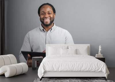 Cheerful African-American young man with dreadlocks holding a digital tablet, testing computer app, is surfing in networks, scrolling news feed in social networks, isolated on grey background Wall mural