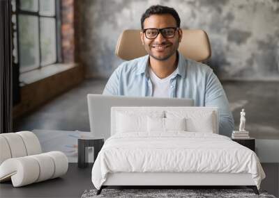 Cheerful indian male student or office worker is using laptop computer sitting at the desk. Friendly and optimistic eastern dark-haired freelancer guy in eyeglasses looks at camera and smiles Wall mural