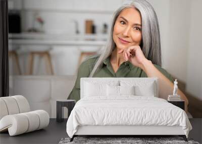 Close-up portrait of charming middle-aged woman with long silver hair sitting in relaxed athmosphere, mature 50s asian female resting at home, looking at camera and smiling with calm smile Wall mural