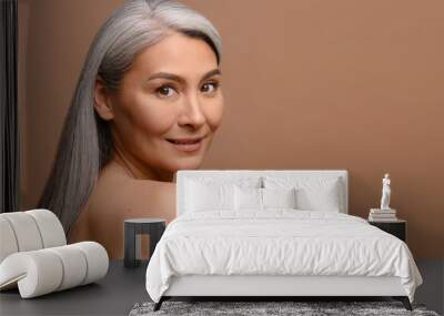 Close-up portrait of fresh and gorgeous middle-aged woman with pure and healthy skin and grey hair, stands with naked shoulders isolated on brown background, mature lady looks at camera and smiles Wall mural