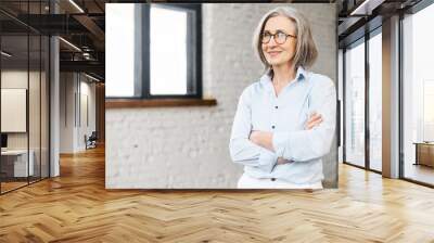 Confident and smiling old senior business woman wearing smart casual shirt and stylish eyeglasses stands with arms crossed in modern office space. A mature bossy lady, elderly purposeful leader Wall mural