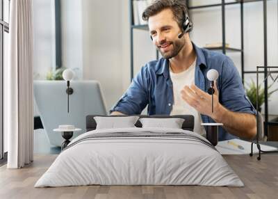 Confident man with handsome stubble, wearing headset, talking in front of computer, wearing casual clothes, talking at computer, discussing work process, gesticulates, chatting with partners, notice Wall mural