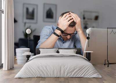 Depressed young businessman holding head in hands, has problem, a laptop on the desk. A guy made a mistake in a work Wall mural