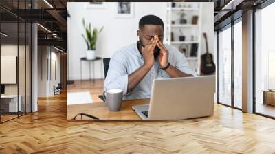 Desperate young African-American business entrepreneur in smart casual attire, touching the face, feeling stressed and overwhelmed while sitting in the office with an open laptop and a cup of coffee Wall mural