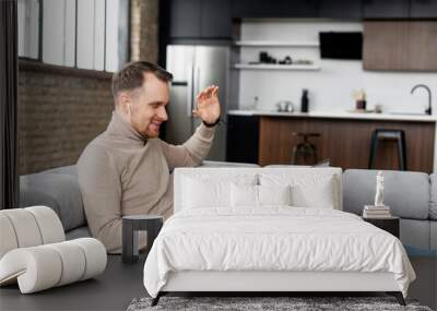 European successful man with earphones greeting by video link, smiling, sitting on a sofa at home, new level of the workplace, telecommunication, with laptop on the lap, meeting with team, waving hand Wall mural