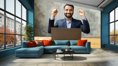 Excited and happy Indian businessman celebrating good deal or win sitting at desk, looks at the laptop screen, male entrepreneur in formal suit raising fists in triumph gesture, business going well Wall mural