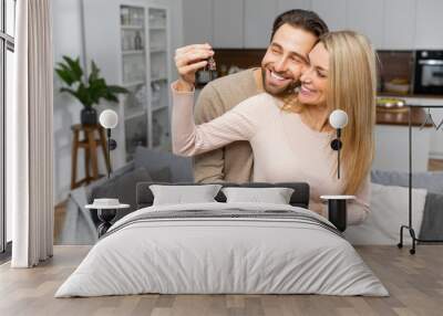 Excited caucasian couple in love move in a new apartment. Cheerful woman in embrace of bearded guy holding the keys, the newlywed look at the camera and smile Wall mural