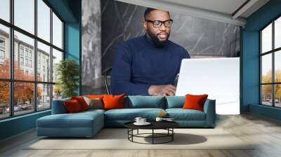 Focused dark skinned businessman wearing stylish eyewear and smart casual turtleneck sits at the desk in contemporary office space, using laptop, conducts business correspondence, answers emails Wall mural