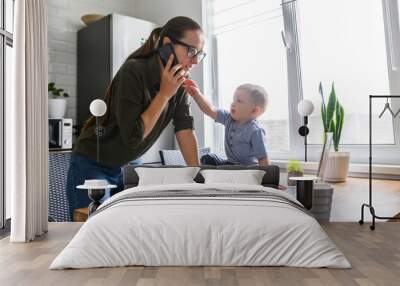 Freelancer mother trying to work from home while babysitting with kid. Mom is talking on the phone while her child is take it out of hand. Wall mural