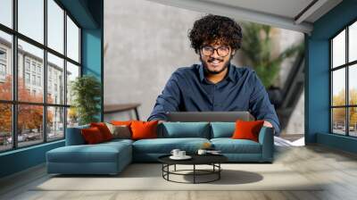 Front view of positive Hindi man in smart casual shirt using laptop while sitting at the desk in his flat. Young Indian male student watching webinars, educational courses, learning on the distance Wall mural