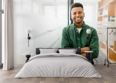 Handsome indian male office employee standing with his arms crossed, looking at the camera and laughs. Portrait of cheerful young man wearing smart casual shirt in modern coworking space Wall mural