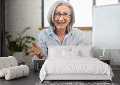 Happy friendly senior female teacher manager with pleasant smile in earphones and glasses looking at camera, sitting at the desk, gesturing and talking online, the view while video calling concept Wall mural