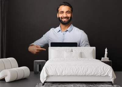 Happy Indian man holding laptop with empty screen and points finger at it, hispanic male employee presenting new computer app, promotion and advertise concept Wall mural