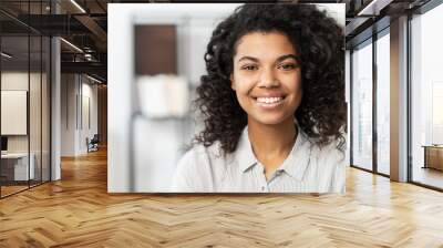 Headshot of a young elegant African American ethnic female with Afro curly hairstyle, beautiful smile and looking at the camera while standing against blurred home or office interior background Wall mural