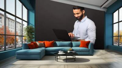 Indian bearded freelancer guy using laptop computer isolated on black background, dark-haired 30s multiracial man in casual shirt typing Wall mural