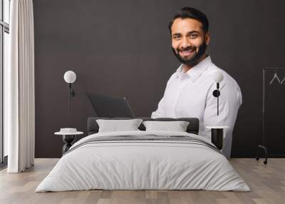 Indian male entrepreneur carrying laptop isolated on black, bearded businessman using laptop, typing, receiving emails, using new computer app, looking at the camera and smiles Wall mural