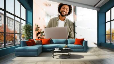 Inspired young african-american businessman looks at camera and smiles cheerfully, standing in modern office and using laptop. Male office employee, freelancer, designer full of new ideas and creative Wall mural