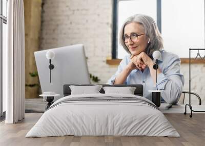 Intelligent elderly gray-haired businesswoman using a laptop in the office. Smart mature woman entrepreneur looks through emails, analyzes tasks, checks report. Senior online teacher conducts lecture Wall mural